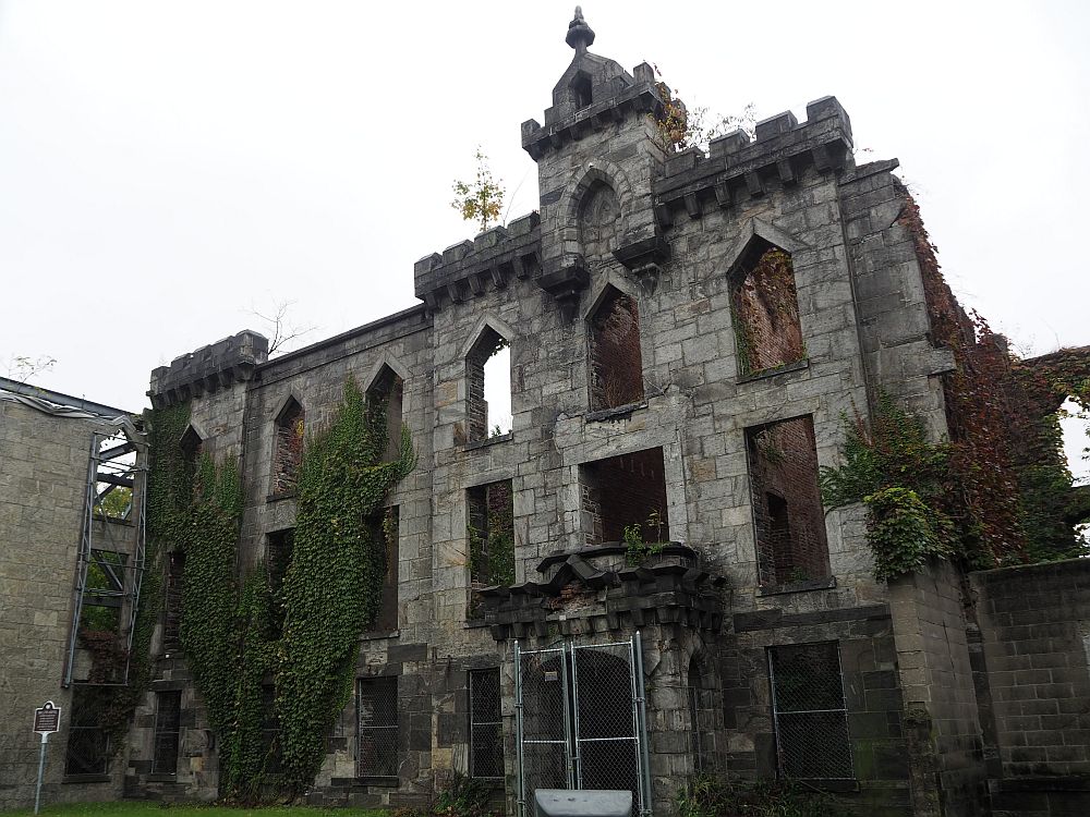 The ruin has an intact front facade but the sky is visible through the windows. Vines grow on the side and small shrubs grow here and there from the stone.