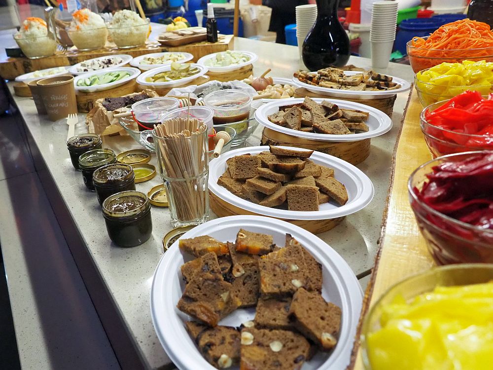 The tasting we got on the Riga food tour: bowls of cabbage on the right: yellow, orange and red. Four plats of assorted slices of bread in the middle. Beyond that, various dips and pickled vegetables on plates. At the back, three bowls of slaw. 