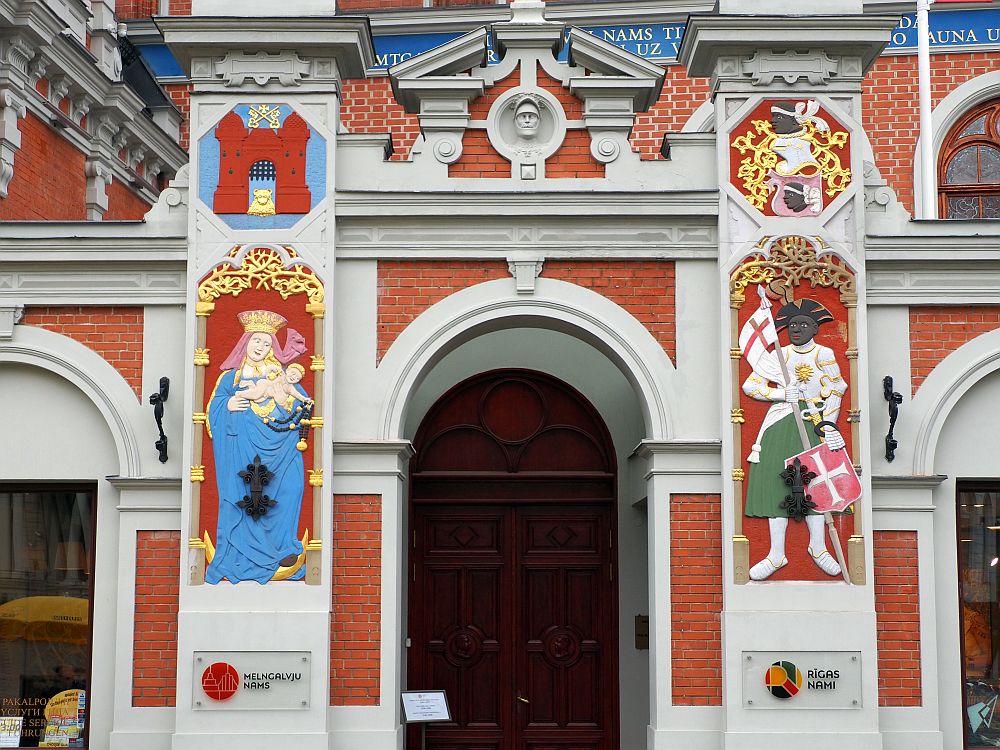 The entryway is arched with a split pediment above it. The large square columns on either side of the entryway have brightly-painted images. The one on the left is a pale-skinned Virgin Mary with a blue robe, a pink headscarf and a gold crown. She is holding a naked baby. On the right is a black man holding a red shield with a white cross and a white flag with a red cross. His pants? skirt? is green and his jacket, with looks vaguely military, is white. 