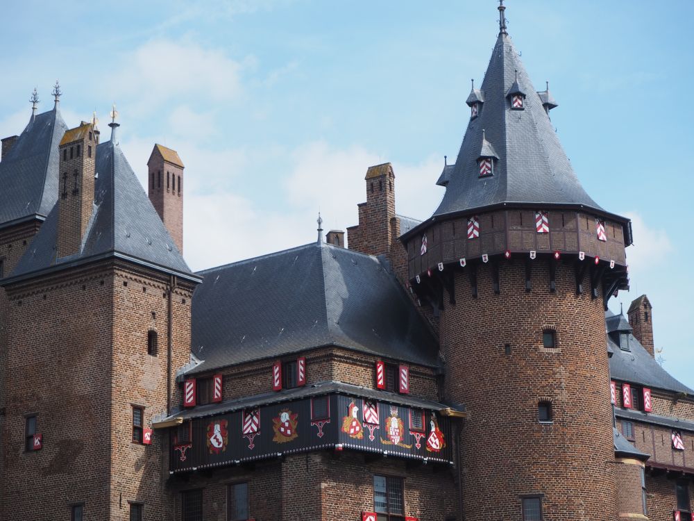 This close-up of the roof line of Castle de Haar shows two turrets. Many of the windows have shutters painted in red and white. A section below the roof has painted images between the windows as well: it looks like coats of arms and cartoon images of knights holding shields. 