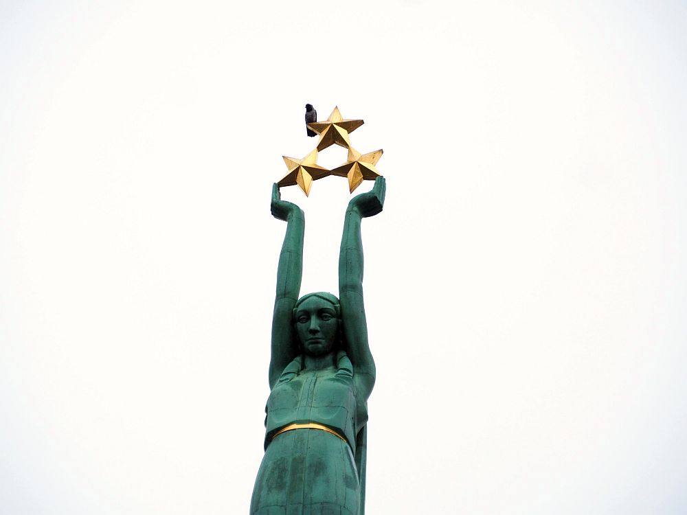 This closepup shows the green woman from the top of the monument from about the thighs up. She looks slightly downward. Her arms are extended straight up and her hands support three 5-pointed gold stars. A bird (a pigeon?) is perched on one of the top star's side points.