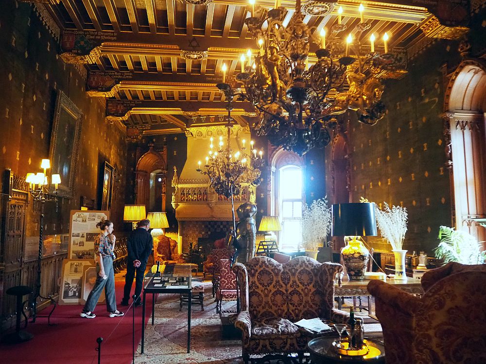 This room Castle de Haar is quite dark, lit by a couple of massive chandeliers as well as light coming in the window. the walpaper is dark red with a small pattern on it. the furniture is set up in clusters of upholstered armchairs and side tables. A suit of armor stands in the center of the room and huge carved fireplace is in the middle of the back wall. 