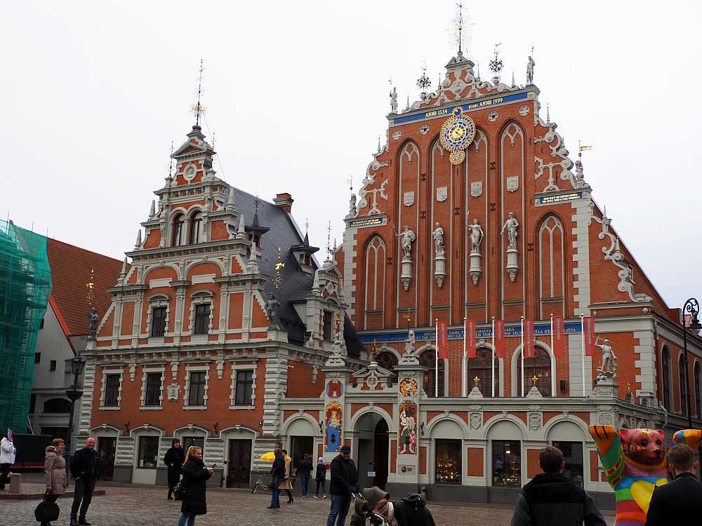 This building looks like two buildings: each with a triangular facade. Both are red brick with white detailing. The building on the right has ornate decorative features between and above all the windows (it's about 4 stories tall). The facade on the right is far more ornate, with a very decorative roof line and a row of human statues across the middle front. The ground floor has arched windows and an arched entryway with colorfully painted figures on either side and statues above it and on the corners of the building.