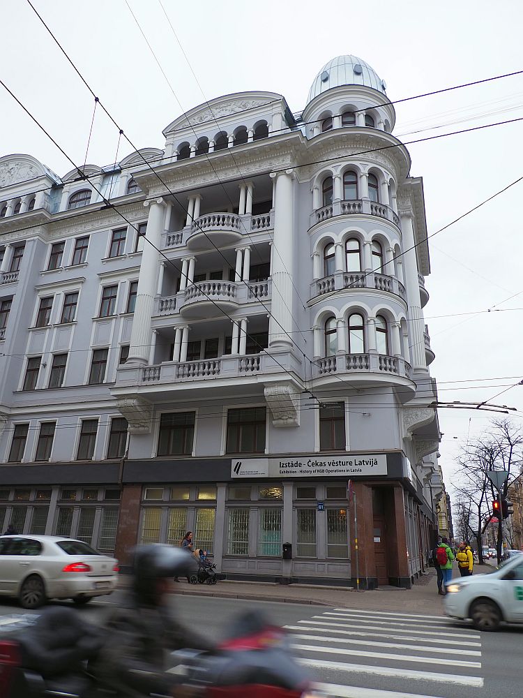 An apartment building on a street corner. It is rounded on the corner and each floor has a half-ring of arched windows on the corner, about 6 stories tall, with a little cupola on top. the sside of the building has half-circular balconies with railings one above the other. The building is light gray with white trim.