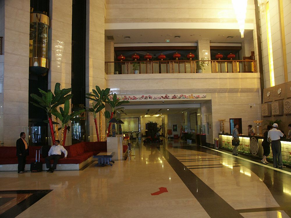 The lobby is several stories tall with a big empty space in the middle with a shiny white floor. On the right are the check-in desks. On the left, a few people sit on a bench. Behind them is a cluster of small trees and behind them, a glass elevater is stopped on the floor above. In the back of the shot a balcony is visible one floor up.