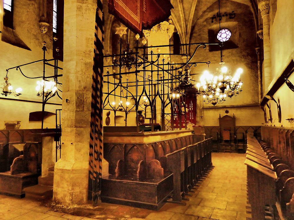 A square wooden structure (the bima) with tall fencework around it in the center of the room. Seats with desks along the side. The ceiling isn't visible in the picture, but it's clear that it's quite high. A bit of the gothic vaulting is visible on the far wall. A chandelier hangs at the far side of the room, and smaller hanging lights surround the bima in the center. 