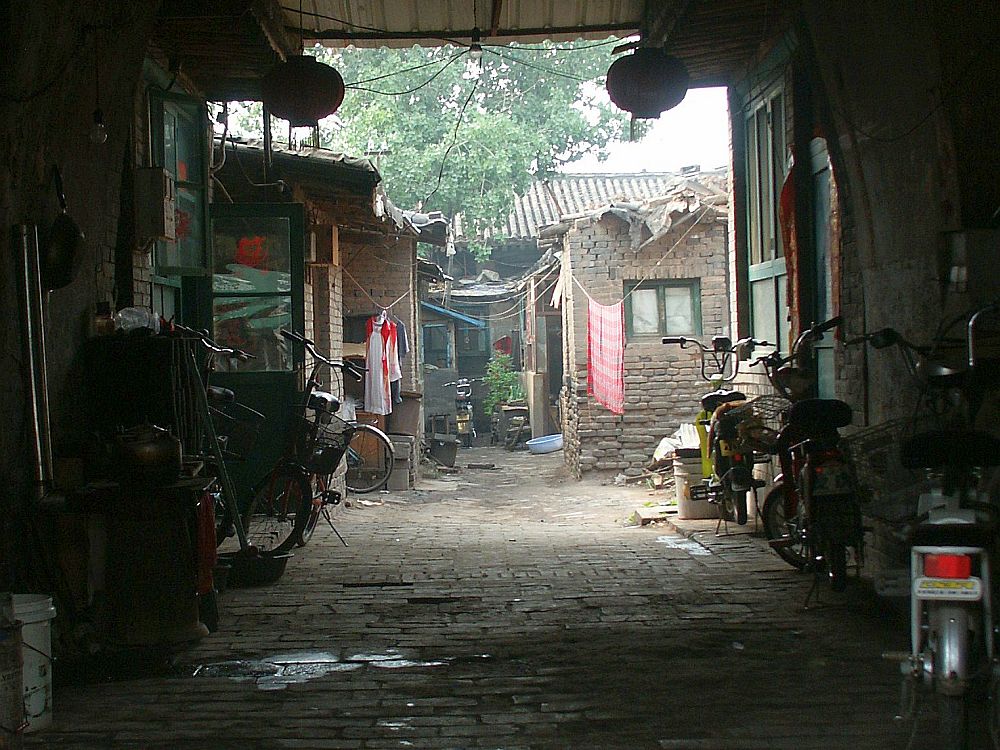 Inside the archway, several bicycles are parked against the side walls. The floor is paved with bricks, wet in places. Beyond the archway is a space that is open to the sky. A row of brick shacks face the center alleyway. Their bricks are rough and their roofs, the bits that are visible, look like they're made of scraps of metal and plastic. Laundry hangs on lines between them.