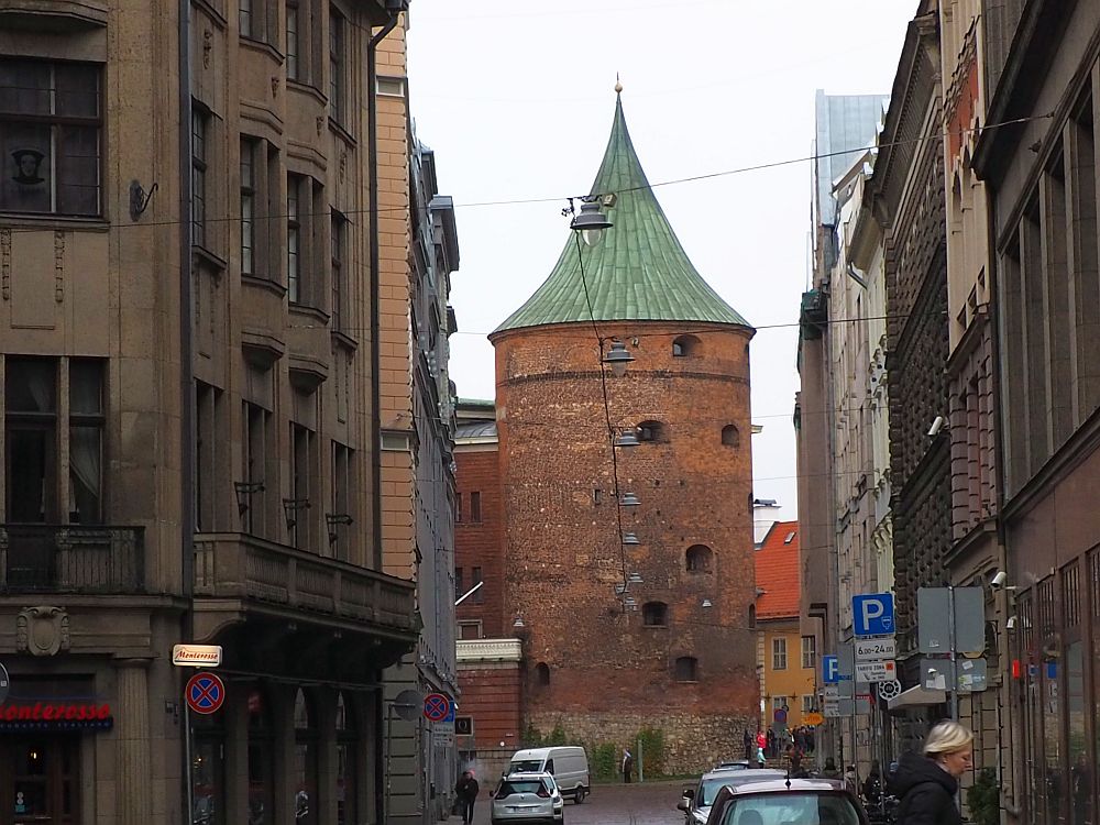 Seen at the end of a short city street, the tower is red-brick, round and several stories high with a green pointed roof. It has only a few small windows.