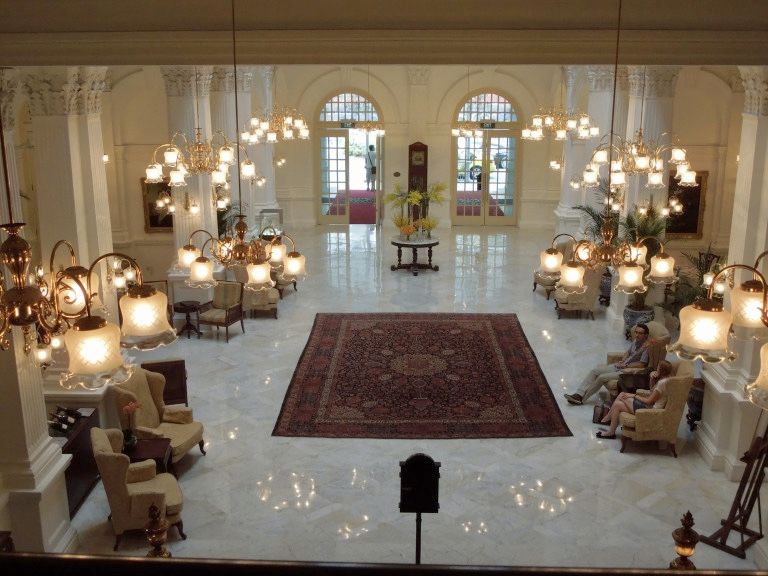 One of the Singapore film locations: the lobby of the Raffles Singapore. A view down from a floor up onto an elegant lobby: white marble floor, chandeliers, armchairs, an oriental rug in the center. 