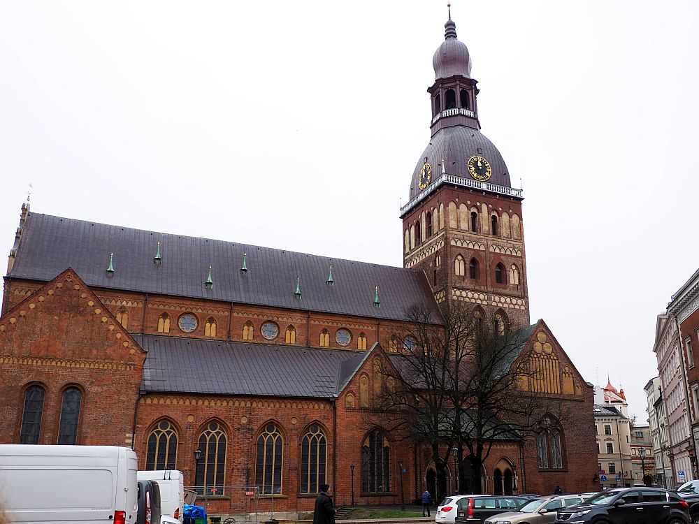 A red-brick church, seen from the side. The side has a row of gothic windows facing on to a parking lot in front. The top of the main part of the church is above that, so a row of round windows is visible above the side roof. ON the right-hand end of the church is its spire, about twice as tall as the building. It is square and has a a few rows of windows. The top of the spire has a dome with a clock and a smaller spire extending up from its middle. That littler spire has a circle of arched windows and above that is a small, elongated dome.