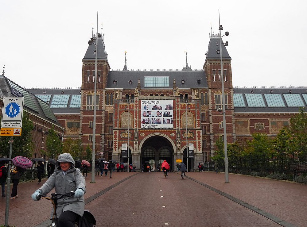 The red-brick museum has a square entrance framed by two turrets. In the center is a large archway, with is the entrance to a bike tunnel through the building. On either side of that is a smaller tunnel for pedestrians. People carry umbrellas on the sidewalk, while people in the bike lane are bundled up against the cold. 