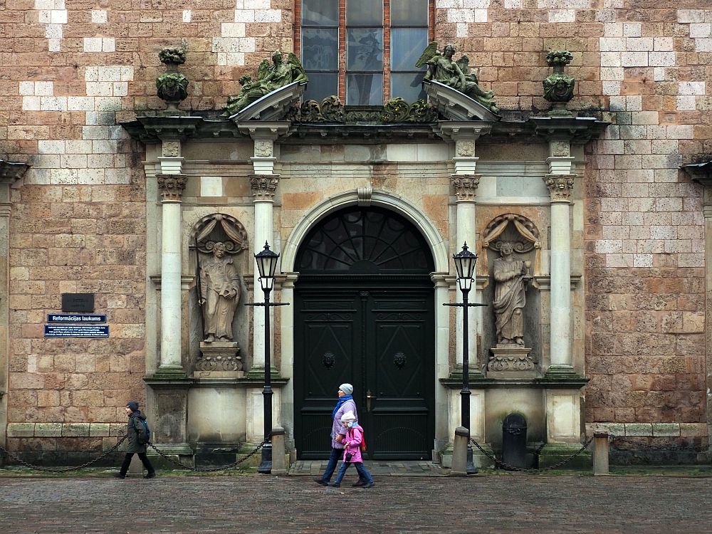 An arched entranceway with a split pediment above it, split to accommodate a large window above. On either side of the entrance is a niche with a statue in it and columns on either side of the statue. A woman and two children walk past the entrance. In terms of height, the woman's head only reaches the bottom of the statues. The wall around the entrance is old stone bricks, in shades of brown.