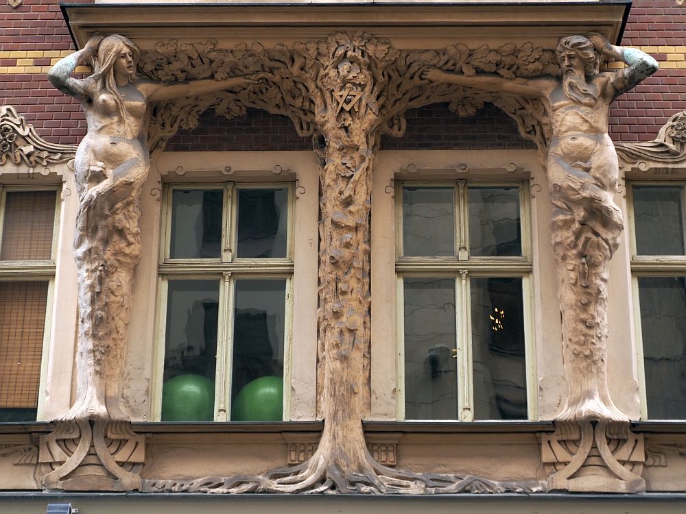 This seems to be an Adam and Eve image. Eve is on the left, with a naked body to the crotch, but below that it looks more like the trunk of a tree. On the right, a male figure with a beard, also naked to the crotch and a tree trunk below that. Between them are two windows, and between the two windows is another trunk image, with roots at the bottom and what looks like vines at the top, that reach to the two human figures on either side. A masonic symbol is also visible in the center tree.