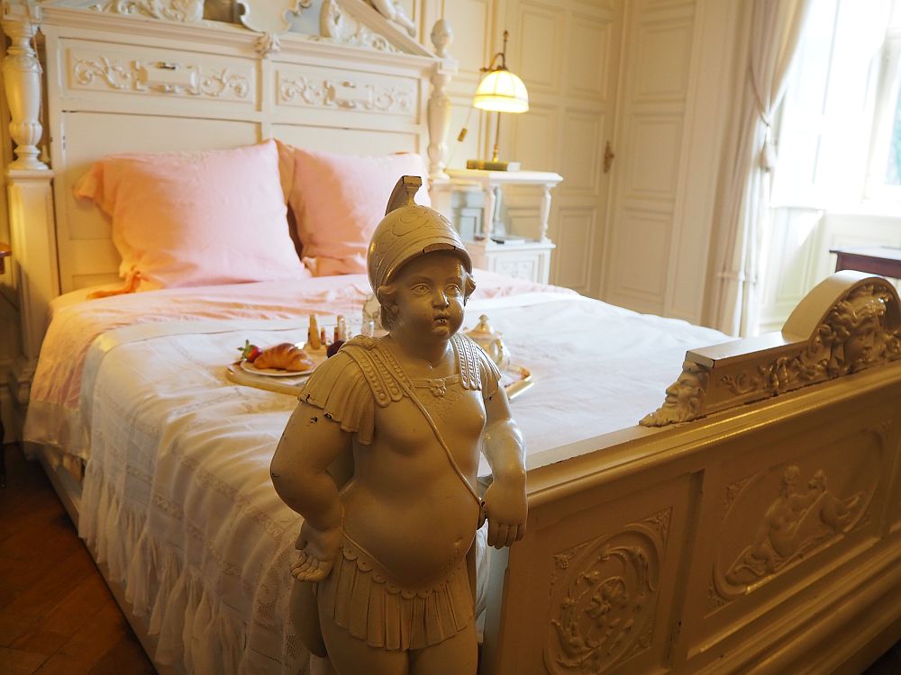 A double bed made with light pink sheets. The headboard is white, carved wood. the foodboard is also carved, but here the carving extends to a complete human, child-sized, facing away from the bed. It is a rather chubby looking child wearing a roman-style helmet and a fringed skirt. 