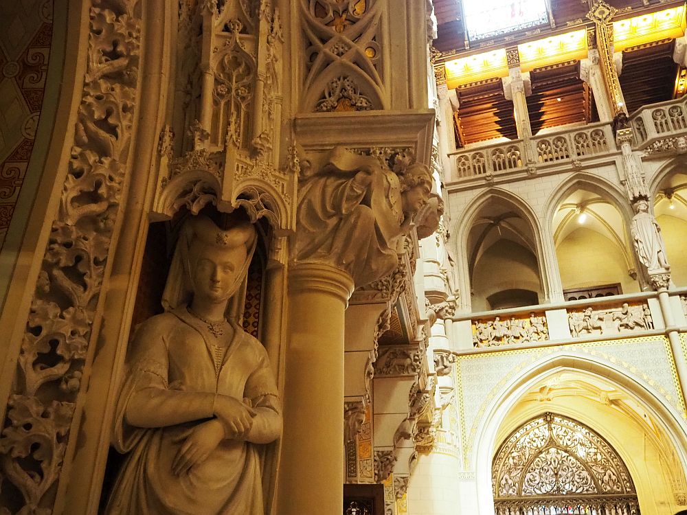 In the foreground, a very complicated carved side of a doorway with an image of a human in front: a woman wearing a medieval headdress and dress. In the background, balconies overlooking the courtyard: each opening has a gothic arch and between the arches are more statues.