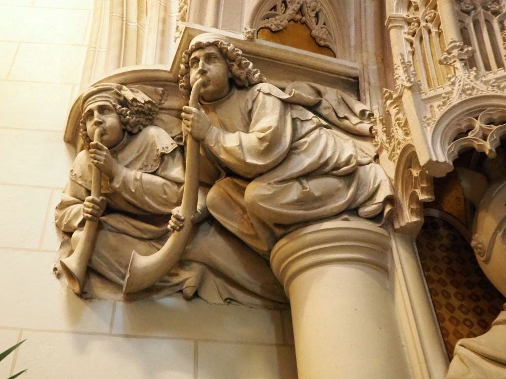 Two carved men, squatting forwards, holding long horns, which they are blowing. The squat on top of a simple column. 