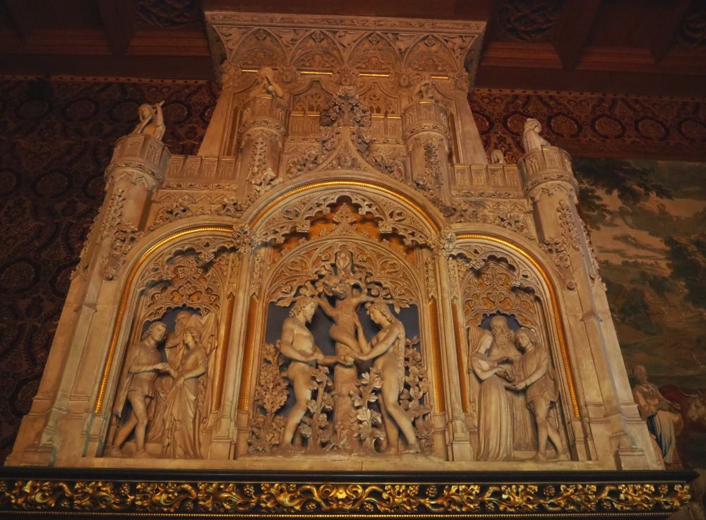 The stone fireplace is carved with three arches. In the middle is an image of Adam and Eve with a tree between them, a snake cured around its trunk. The images in the two outer arches other scenes but I can't tell what they're meant to represent. They both have figures who are dressed in medieval style. Above the arches rae more carvings, mostly flowers and leaves. 