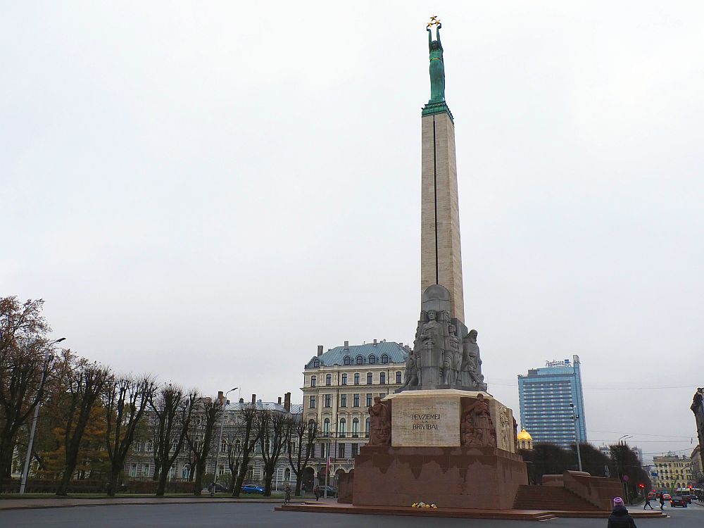 A very tall and think monument against a grey sky. The base in red stone with sculptures around the base. On the top of the base is a cluster of human figures in a grey stone. Above that is a tall, square pillar that narrows toward the top. At the top is a figure of a woman in a green tone. Her arms are extended straight above her head, holding a cluster of 3 stars in gold. 