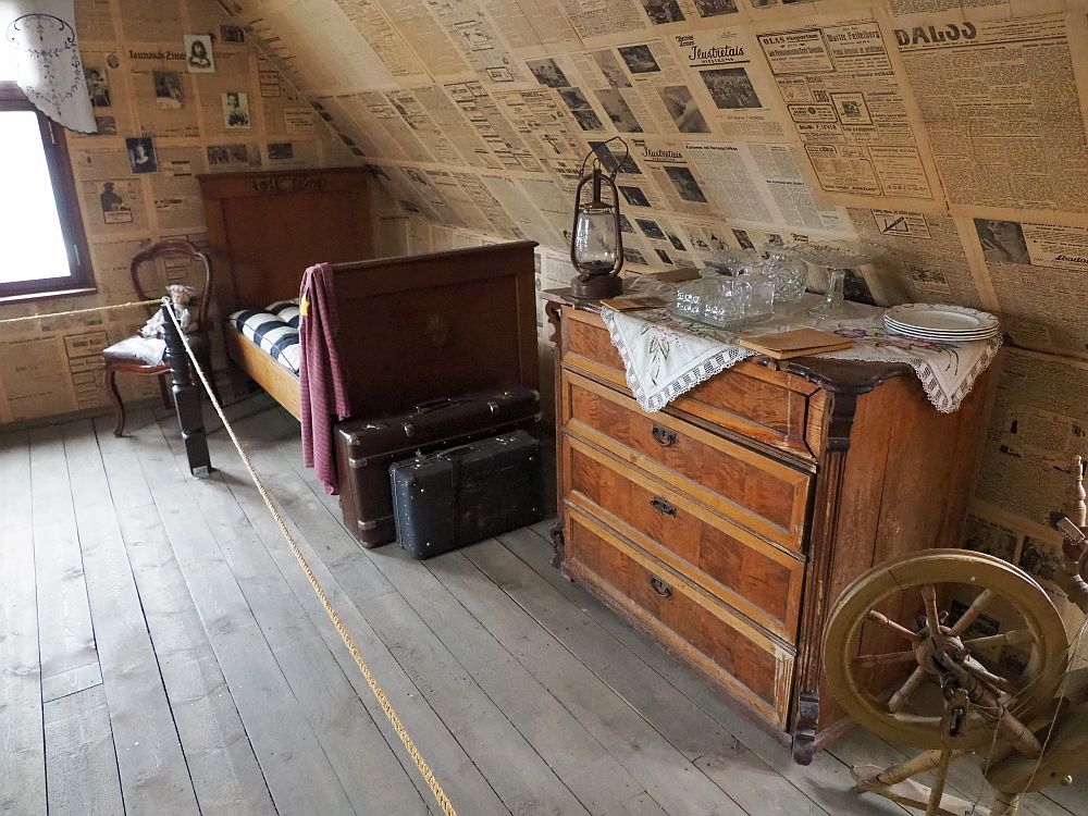 The wall is slanted under the roof and wallpapered with newspaper. A bureau with a few objects on it: glasses, a lantern, some plates. A spinning wheel is partially in view in front of that. Beyond the bureau is a single bed and at its foot, two suitcases. Next to the bed is a chair with a doll sitting on it. 