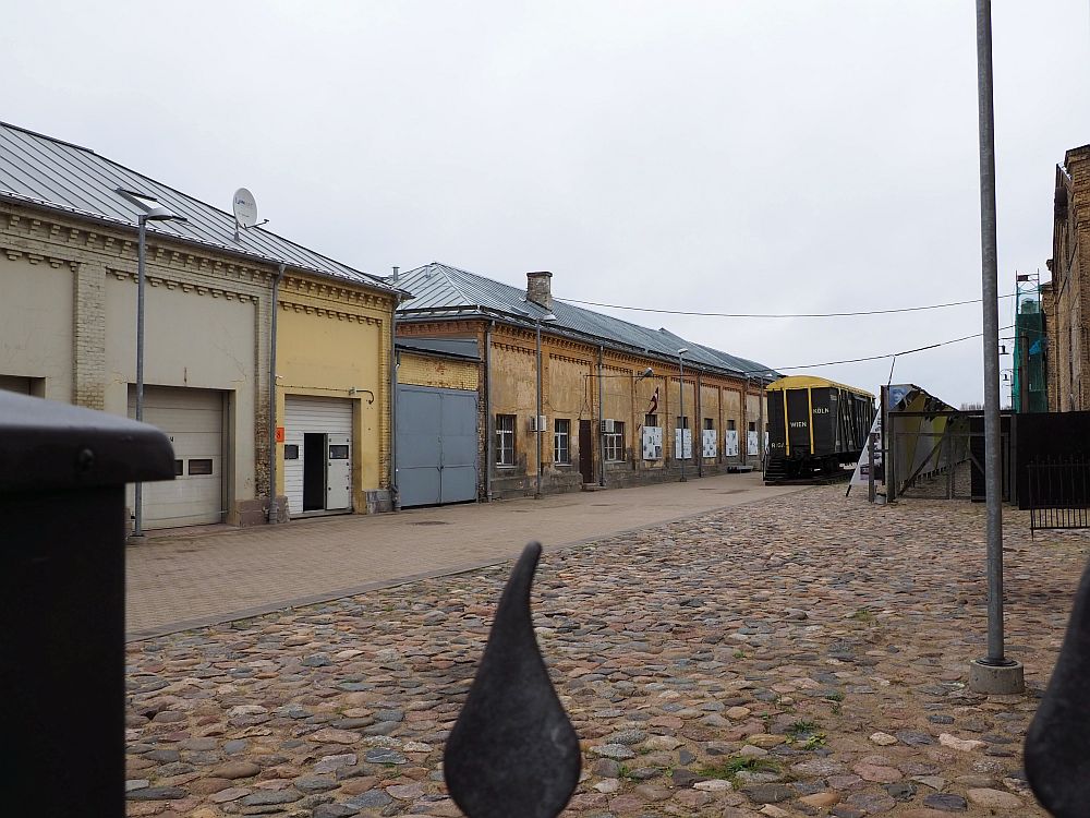 A cobbled courtyard in front with a row of abandoned-looking buildings on the left. Looking down the center a box car is visible in the distance, dark green. 