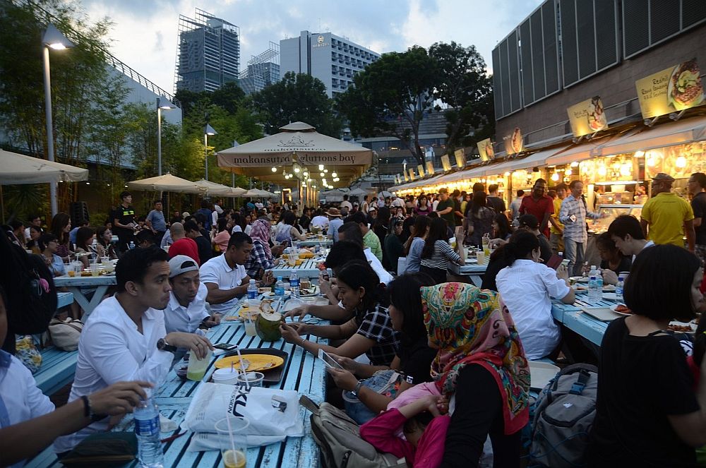 Outdoor, clearly at dusk, long lines of picnic tables, which all look completely filled up with people eating. To the right is a row of food stalls, lit brightly, and people stand waiting for their food. 