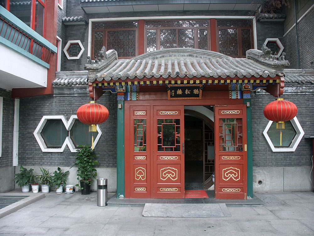 The entry is four doors wide, all painted red. A small roof juts out above it, curved in the traditional Chinese way. A red paper lamp hangs on both corners of the little roof. On each side of the entrance are six-sided windows: two on the left and one on the right. 