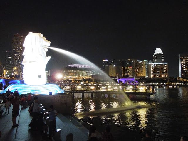 The Merlion statue is on the left of the picture, brightly lit up, as is the water spewing from its mouth to the right, into the river. It's nighttime, and tall buildings are in the background, with white lights in some of the windows against a black sky.