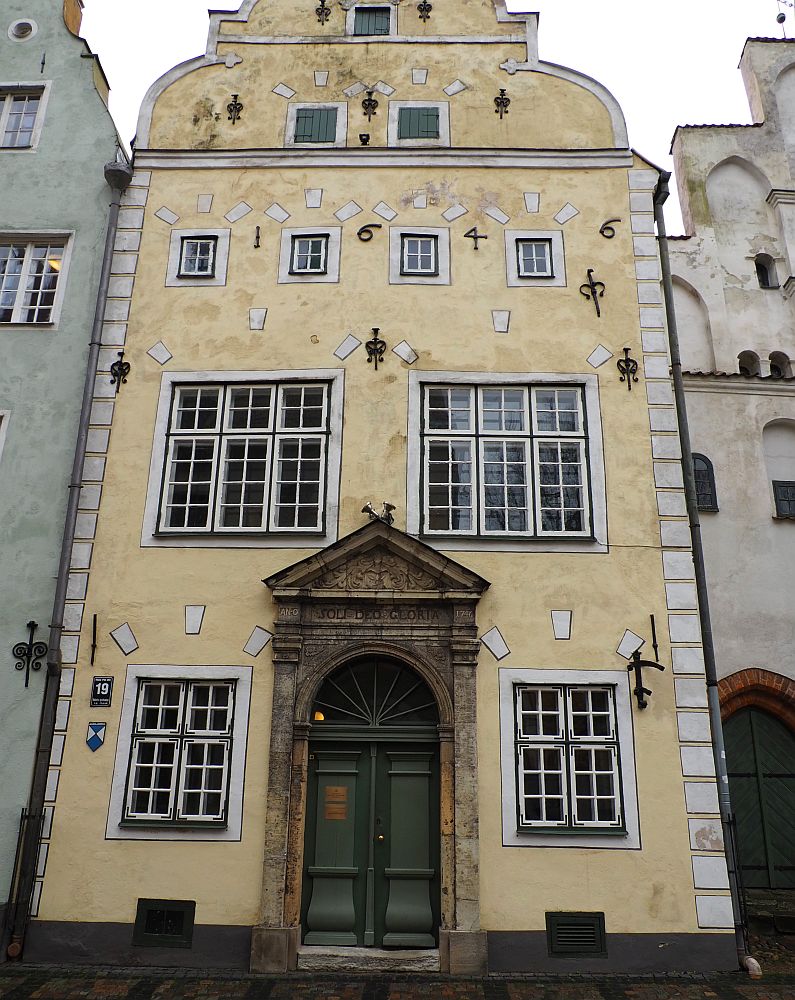 The picture is mostly filled with the middle house. It is pale yellow with an arched doorway with a triangular pediment above it. The windows of the building (3-4 stories) are multi-paned and edged in white and black. Letters on the 3rd story read "1646."