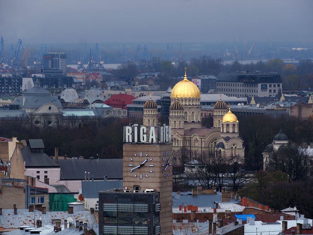 In front, a square tower with no windows but with huge signs on the two visible tops reading RIGA and, below that on the two visible sides are huge clocks. Behind that is the Russian Orthodox Church with its gold domes. In the evening light the domes seem to shine. Beyond the church is a blurry view of the city.