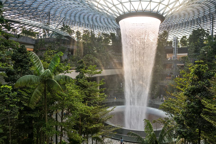 In a massive round hall in Changi Airport, the ceiling is a network of metal holding glass panels. In the center, the water falls in a cylindrical shape through a round hole in the roof. Trees grow all around it, but here and there some of the balconies are visible beyond the waterfall. A few people are barely visible at the bottom of the picture, where the waterfall falls into a big round opening. 