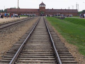 The main entryway to Auschwitz as viewed down a railway track. Low brick building with a watchtower over the entryway in the center.