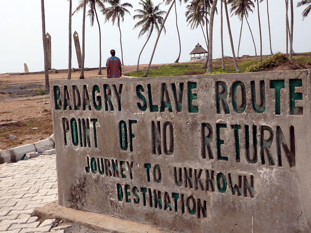In the foreground, a concrete sign carved with capital letters reading: BADAGRY SLAVE ROUTE
POINT OF NO RETURN
JOURNEY TO UNKNOWN
DESTINATION
Beyond the sign is a flat piece of land, partly brown dirt and partly grass-covered, with a few tall palm trees spaced over it. Beyond them is the monument, just two concrete poles, slightly leaning inward, each with a round hole at its tip. A man walks toward the monument; only his upper body is visible above the sign.