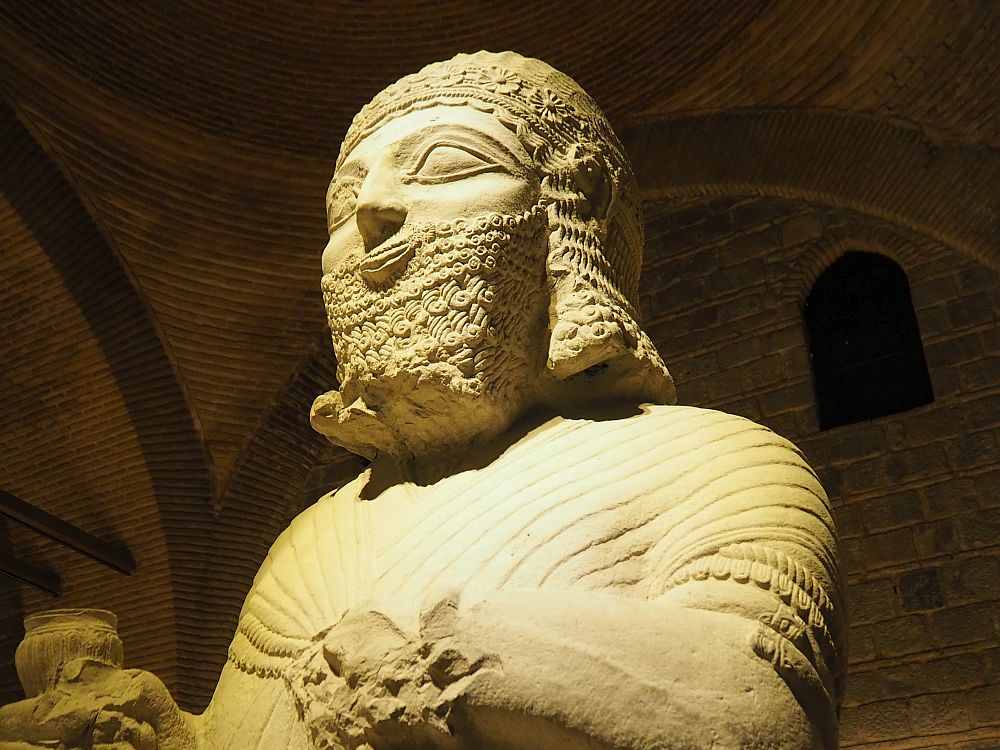 The photo only shows the head and chest of the statue. It has detailed facial hair: the beard is square and carved in neat curls. So is the hair, and a decorative hat is carved with flowers and what looks like decorative lace. The chest is simpler, carved as if he is wearing a cloak that pins in the center of his chest. 