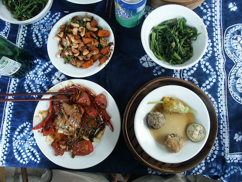 Four dishes, seen from above, on a blue and white batik tablecloth. Top left, chicken with cashew nuts. Top right: some sort of greens. Bottom left, beef, I think, with red peppers and tomatoes. Bottom right: three small dumplings, all different, and a fourth one that is a stuffed flower. 