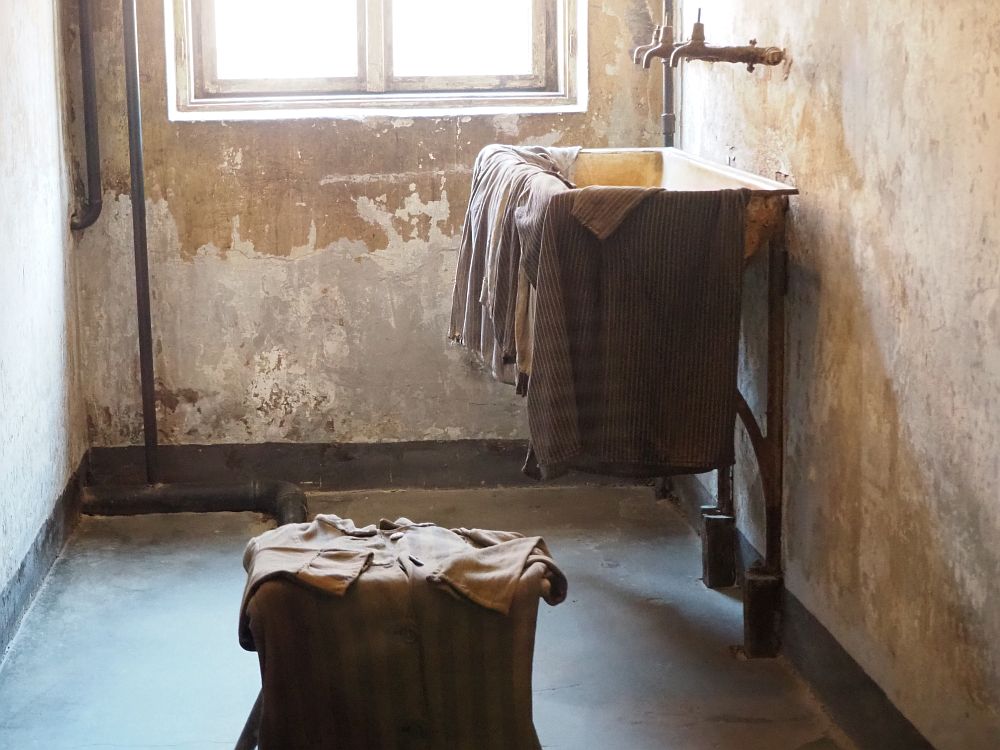 A low stool stands in the center with some clothing laid on it. On the right is a sink with faucets above it. Clothing is draped over the edge of the sink. The room is quite small and the walls are various shades of brown and white.