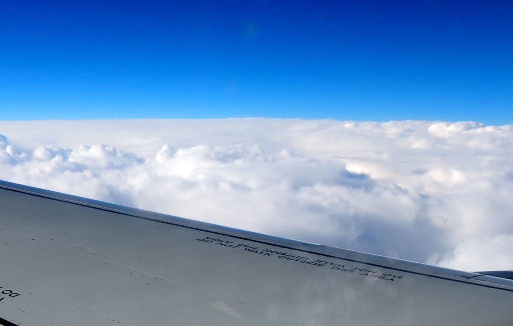 A view over an airplane wing onto fluffy white clouds. Above them, brilliant blue sky.