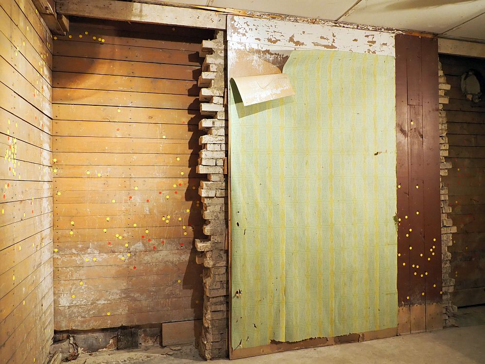 The wall has visible layers. About a third of it is horizontal wooden boards on the left. The middle half is brick in front of the wooden layer, so the brick wall ends with an uneven vertical cut. In front of the brick is another layer - wood or drywall or something, that has peeling, light green wallpaper on it. On the right is a small section of more wood, this time on top of the brick rather than behind it. Here the wood boards are vertical. All over both wood layers are little yellow and some red dots. 