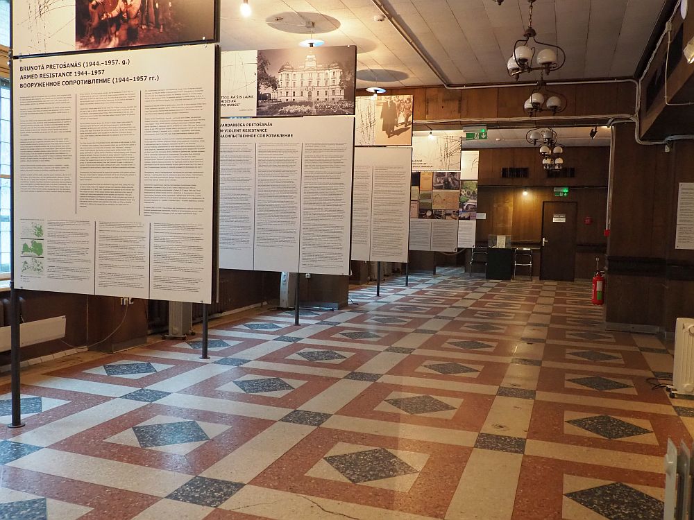 A large room, the floor in neat tile squares. Along the left, where the windows are, stand a series of 5 large sign, perpendicular to the left wall. Each is printed with some of the history with a photo at the top. The history is written three times: in Latvian, in English and in Russian.