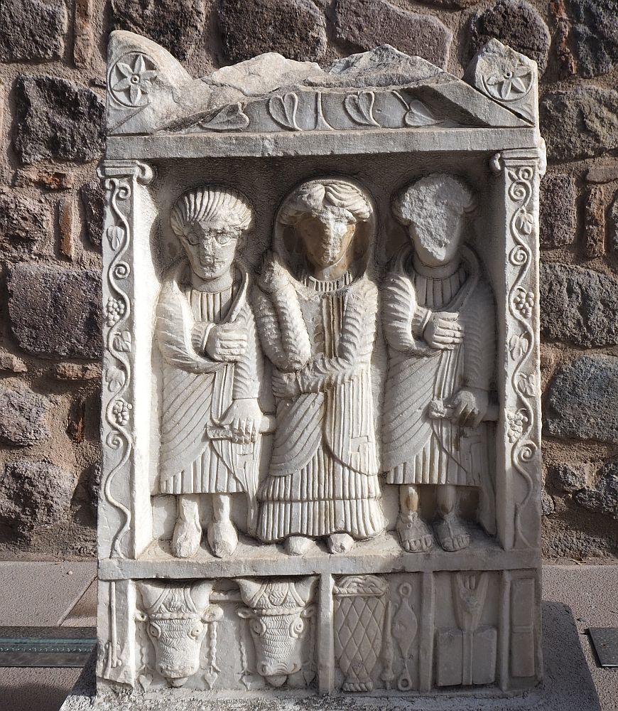 The grave marker is carved so that three human figures stand in a framed niche. The one in the center may be a woman: the robe is longer and he/she wears a hood as well. The ones on either side each hold something in their left hands - a scroll, perhaps? - nd have their right hands in a sort of sling. The faces are eroded or damaged. Around the frame grape leaves are carved. Below the feet of the three figures are smaller frames. The one on the left has the faces of two cows or oxen. The one in the middle shows a basket or perhaps a fish trap. And the one on the right isn't clear. 