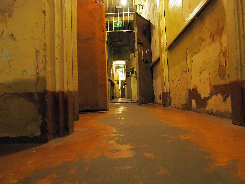 Looking down a hallway, in the middle distance is a door, open, with a heavy lock and bars above it to the ceiling. Beyond that are more open doors on the right with heavy locks. The paint has worn off the floor except on the edges. Along the right-hand wall of the hallyway is a horizontal bar. 