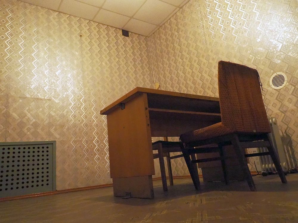 Photo taken from ground level, looking upwards. Two walls of the room are visible, covered with ugly wallpaper in a zigzag design in shades of gray. A wooden table stands in the middle with a straight chair on either side. 