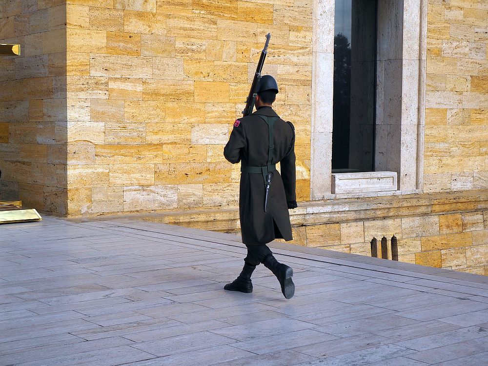 The guard is walking away and to the left. He wears a black trenchcoat, black pants, black socks and black shoes and a black hat too. He has a knife hanging from his belt and carries a long with his left hand, leaning the gun on his shoulder, pointing upwards. 