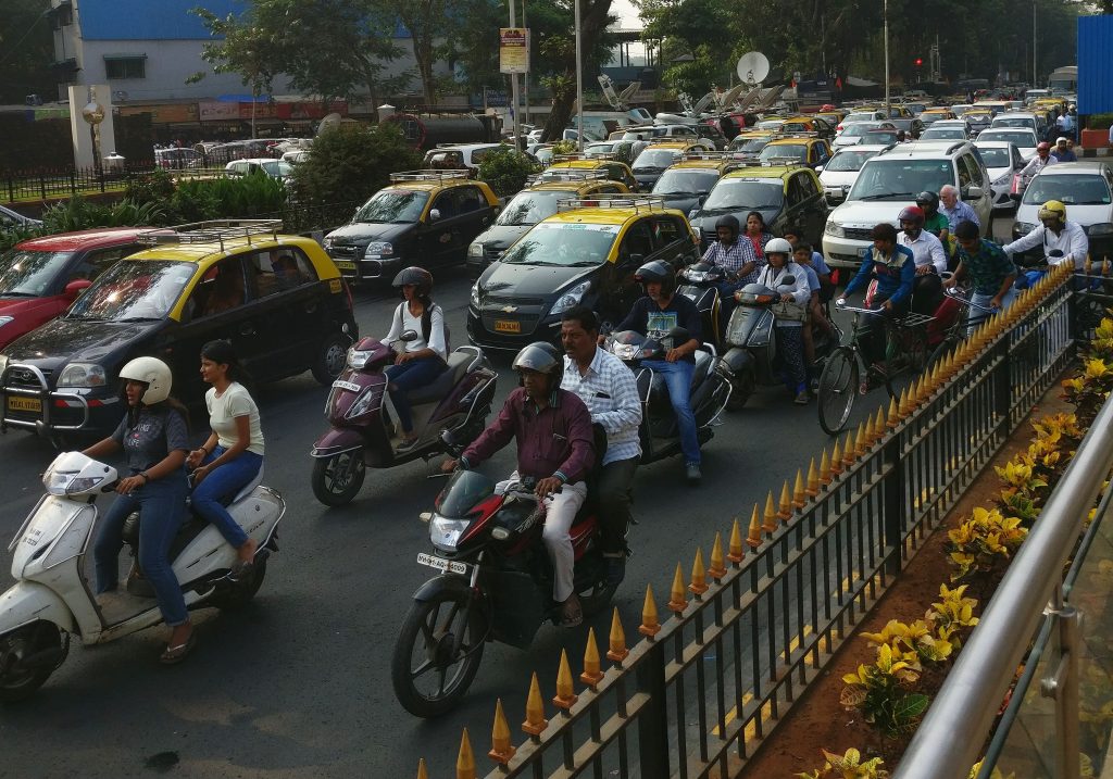 A very full roadway, with, nearby, a group of motorcycles, some carryign two people. Only some of them wear helmets. The rest of the traffic appears to be cars and taxis, and they're not moving, judging by the fact that the motorcyclists in the foreground have their feet steadying their motorcycles. 