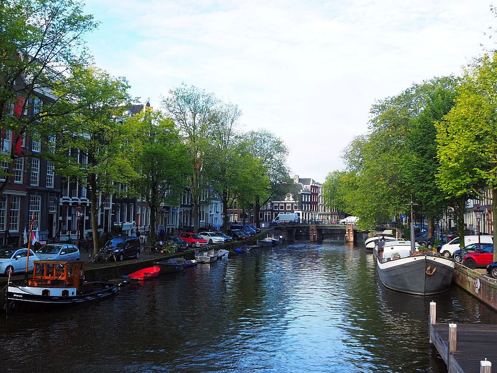 A view down a canal. Row houses on either side, boats moored along the side, a bridge crossing it in the distance.