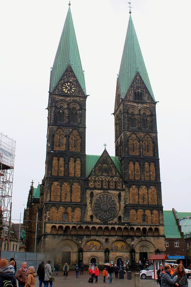 The St. Petri Dom has two very tall spires, both square with green pointed roofs. The building is red brick with archways across the whole ground floor and a rose window centered above the entrance and between the towers.