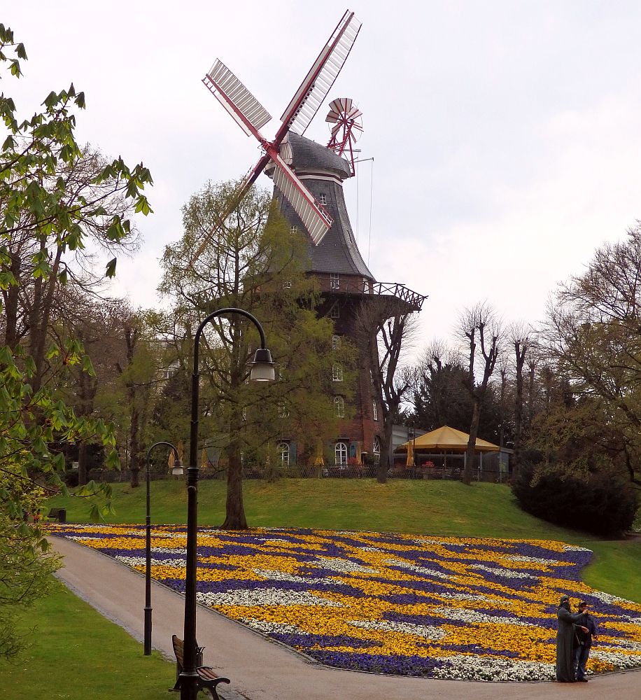 The windmill is quite high: it looks like the cylindrical building under it is four stories tall, made of red brick. Above that is the circular balcony and above that the windmill itself, with 4 vanes and a smaller windmill on the opposite side. In the foreground are colorful flower gardens in yellow, purple and white. 