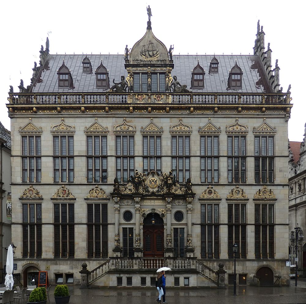 A grand facade: grey bricks with ornate decorations above each window, around the roof line and especially over the central doorway. The building is symmetrical, with the doorway in the center, three windows on either side on the ground floor and 9 windows on the upper floor. There are small dormer windows in the roof and a big one centered with a point above it holding a figure of a man. a stairway leads up to the front door from each side. 