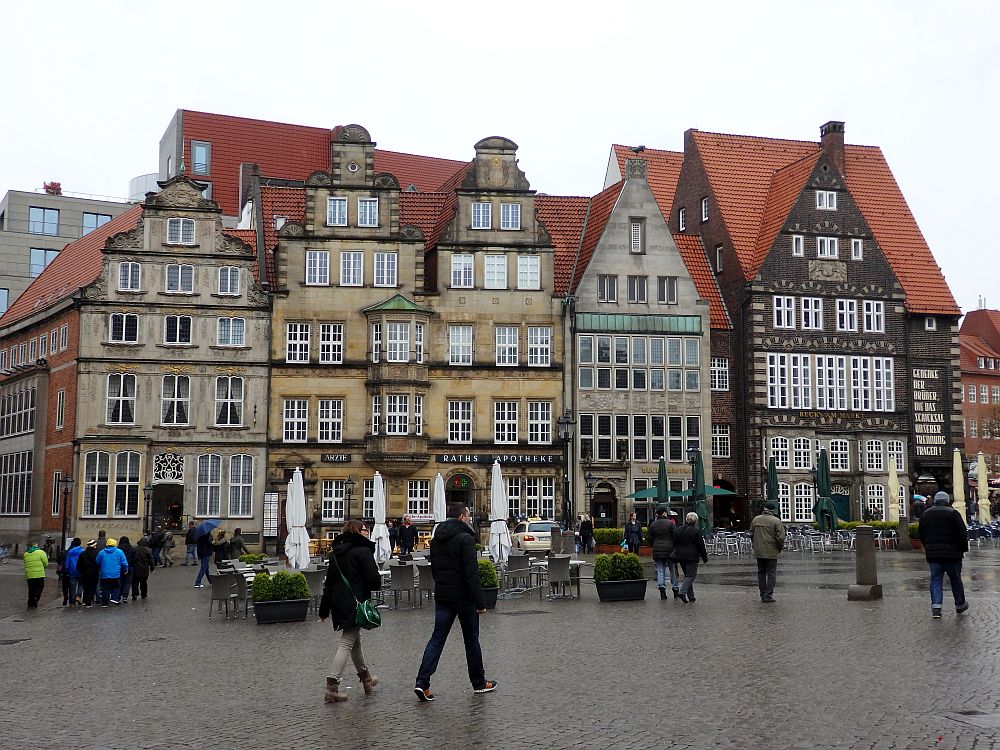 A row of buildings, all in shades of brown with red roofs. Some have stepped roofs, some simpler points. All have a lot of windows. In front of them are tables and chairs for an outdoor cafe, but it's rainy and no one is sitting there. People are walking by.