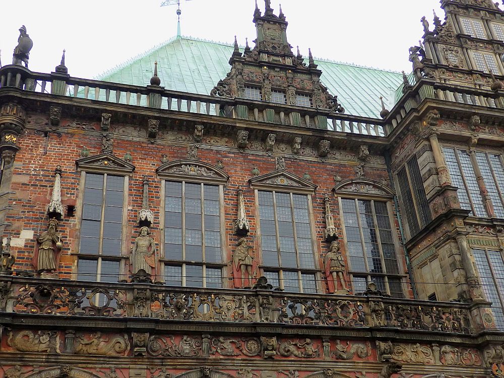 The building is red brick. The balcony has an ornately carved railing and the wall behind the balcony has a row of tall paned windows with statues between each two.