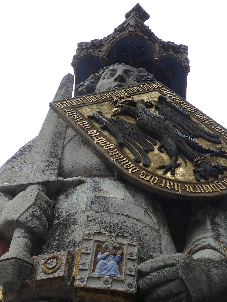 The statue is seen from below in this photo. The figure of a man (I think) holds a sword in one hand and has a shield across his chest showing a double-headed eagle on a gold background with gold words around it. His belt buckle portrays a woman (I think) ina blue robe, playing a lute or mandolin or similar.